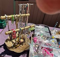 Overview of necklaces and bracelets hanging on two-tier gold-colored stand with various jewelry findings and accessories in plastic bags nearby