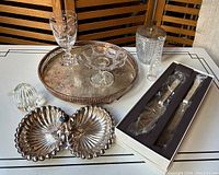 Full overview of lot with all items arranged on table: Waterford bud vase, pattern glass compote, silverplate footed tray, Vera Wang boxed cake set, mid-century chrome nut dish, crystal paperweight, silverplate lidded jar, Gorham crystal goblet.