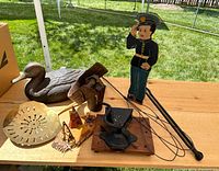 Photo showing the entire collection on a wooden table outside, including the carved wooden duck decoy, wooden officer statue, iron tongs, rug beater, bell collar, brass roaster, wall grinder and souvenir village figures.