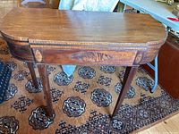 Full view of the oval-shaped antique game table showing wood grain top, apron inlay, and tapered legs.