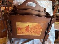 Frontal angled view of the wooden magazine stand showing three compartments and the hunt scene artwork on the front panel with noticeable peeling.
