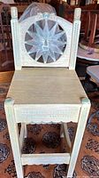 Front view of the hand painted wooden Mexican stool showing the carved sun face on the backrest, scalloped apron, and turned wood posts with painted details.