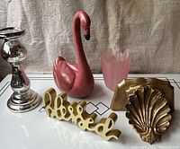 Five home decor items on a white table surface with beige textured fabric background, including a pink wooden swan, pink glass vase, gold 'blessed' word decor, gold Florentine shell shelf, and mercury glass candlestick holder.
