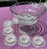Clear glass punch bowl on pedestal base surrounded by six cups on matching saucers on purple cloth.