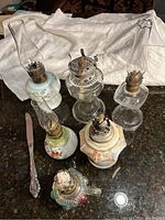 Wide shot showing six different small oil lamps arranged on a dark surface with white cloth in background. Includes a butter spreader (not included) for size reference.