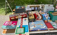 Overhead photo showing multiple vintage toys including games, dolls, and educational sets arranged on a table with focus on Koo Zoo game, Monopoly, and Springfield Railroad game boxes.