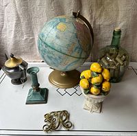 Photo of the lot items including vintage globe, antique teapot, brass candlestick, pottery lemons, large green bottle with corks, and brass trivet on a white table.
