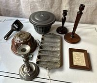 Photo of all items arranged on white table surface showing copper fish mold, black cornbread pan with lid, two wooden candlesticks, pewter candlestick, rectangular mold pan, and framed paper cut art.