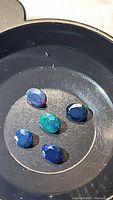 Five natural Ethiopian black opal faceted stones in oval and round cuts placed in a shallow black dish under natural light, showing vibrant play-of-color.