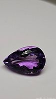 Close-up photo of a pear cut purple amethyst gemstone showing facets and rich color on a plain background.