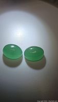 Pair of polished green onyx oval cabochon gemstones shown from top angle with light reflection highlighting smooth surface and translucent green color.