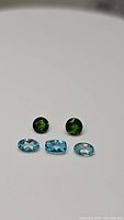 Five natural gemstones displayed on a white background, three blue apatite and two green chrome diopside.
