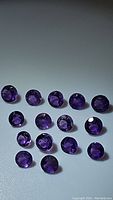 Photo showing 15 round calibrated deep purple Brazilian amethyst gemstones laid out on a flat surface, displaying faceted cuts and clarity.