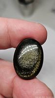 Close-up photo of oval polished Gold Sheen Obsidian held between fingers, showing gold reflective flash on dark base.