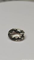 Photo of a single oval cut green amethyst (prasiolite) gemstone with light teal green color from one angle. Multiple facets visible, clear and transparent with light reflections.