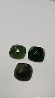 Three fancy cut green jade gemstones placed on a white background, showing dark green grey hues and faceted surfaces.