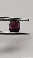 Clear close-up photo showing the deep reddish-purple cushion cut spinel held by tweezers against a white background, highlighting cut and clarity.