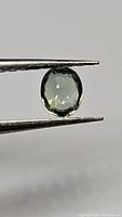 Close-up of bright green oval cut natural sapphire held by tweezers showing facets and color.