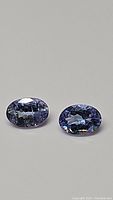 Pair of oval cut bluish-purple tanzanite gemstones with visible rainbow inclusions shown against a gray background.