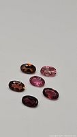 Six oval cut natural tourmaline gemstones in various shades of pink and some brownish tones displayed on a flat surface.