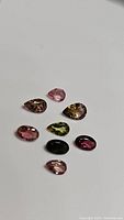 Group of eight natural tourmaline gemstones in pear and oval cuts, arranged on white background, showing variety of colors and clarity.