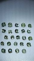 Top-down view showing 24 loose natural alexandrite round cut gemstones arranged in a grid on a textured white background. Varied green and yellow shades visible under lighting.