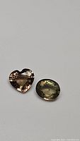 Two natural tourmaline gemstones showing a heart-shaped peach pink stone and an oval-shaped champagne colored stone on a neutral background.