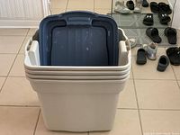 Four stacked Rubbermaid RoughTote storage bins in light gray with matching lids, placed on a tiled floor.