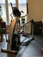Side view of the elliptical trainer showing the frame, pedals, and handles positioned on a black tiled floor near windows and plants.