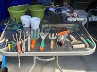 Photo showing assorted hand gardening tools including hand snips, hand rakes, bulb planter, seed holer, hand hoe, weed pick on a glass table next to plastic pots and gardening accessories.