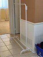 White metal clothing rack standing on tiled floor near a beige wall and door, showing overall structure and rectangular base.