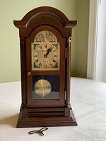 Full view of mini grandfather clock showing wooden casing, arched top, decorative columns, glass panels, pendulum, and winding key on flat surface in front.