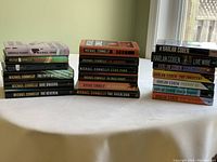 Photo showing 3 stacks of paperback and hardcover novels by Michael Connelly and Harlan Coben on a white surface against a green wall with window light.