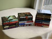 Three stacks of paperback and hardcover novels by authors starting with 'R', shown on a cream colored surface near window.