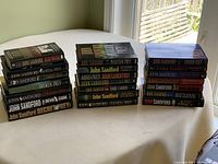 Three stacks of John Sandford novels totaling 21 books on a table near a window under daylight.