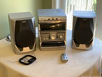 Sony stereo main unit with dual cassette decks, 3-disc CD changer, control buttons, and large volume knob, flanked by two matching speakers. Power cable visible.