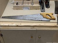 Hand saw with light-colored wooden handle and blade on a work surface with mitre boxes in background
