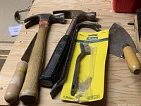 Various hand tools including hammers, a wire brush, metal scrapers, garden trowel and caulking dispensers laid out on a wooden surface.