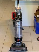 Full view of upright Hoover vacuum cleaner with clear dirt bin and red cleaning brush attached on side.