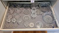 View of multiple crystal vases, small plates, bowls, and dishes arranged in a drawer on a gray liner. Includes several small round or square dishes, small glasses, and decorative pieces.