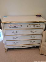 Front view of white wooden dresser with gold trim, showing two smaller top drawers and three larger bottom drawers with decorative handles.