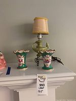 Front view of lamp and vases on shelf, showing lamp with beige shade and green ceramic base, and two vases with floral patterns.