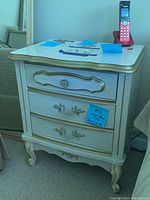 Photo of one nightstand with three drawers, white finish, gold trim and decorative handles, damaged drawer visible, contents on top not included.