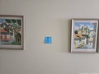Two framed paintings hanging side by side on a light-colored wall depicting Spanish village scenes.