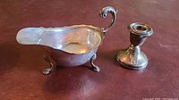 Sterling silver sauce boat with ornate handle and feet, showing tarnish and reflections, placed on brown leather surface.
