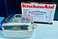 KitchenAid branded stainless steel roaster with lid shown with original box in the background on blue surface.