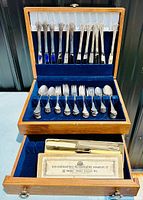 Opened wooden chest revealing silverplated flatware including forks, knives, and spoons neatly arranged in blue velvet slots.
