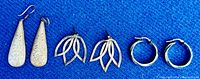 Three pairs of sterling silver earrings on blue textile, includes elongated engraved drop earrings, stylized leaf drop earrings, and plain hoop earrings