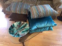 Six cushions stacked on and around a glass table with a large decorative bowl in front. Shows variety of cushion fabrics and colors including striped and solid.
