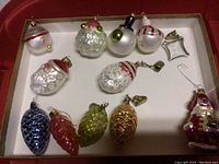 Box with assorted glass Christmas ornaments including Santa Claus heads, round balls, bell shaped ornament, and pine cone shapes.